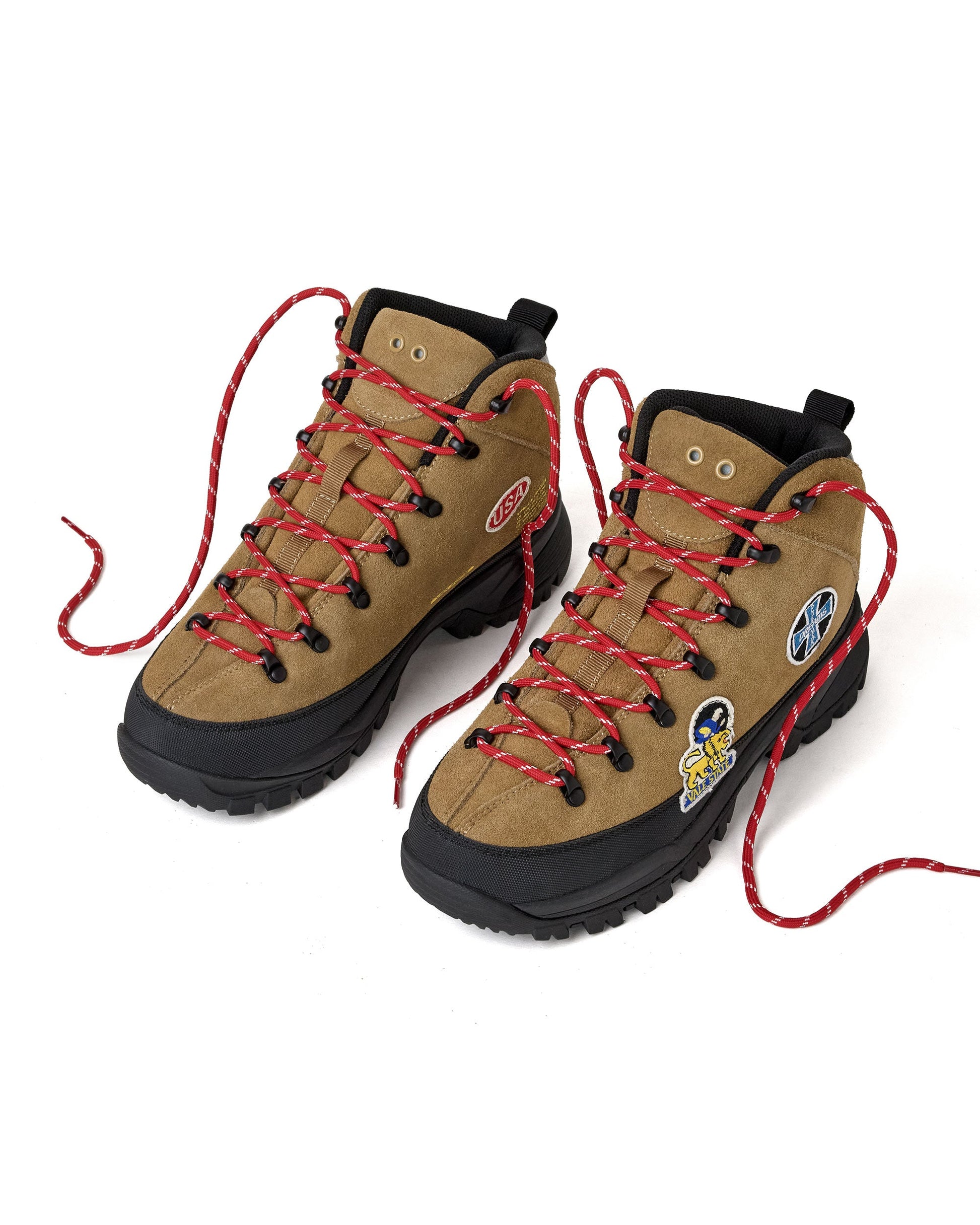 A pair of tan suede high-top hiking boots with red-and-white laces, black soles, and various colorful patches on the sides.