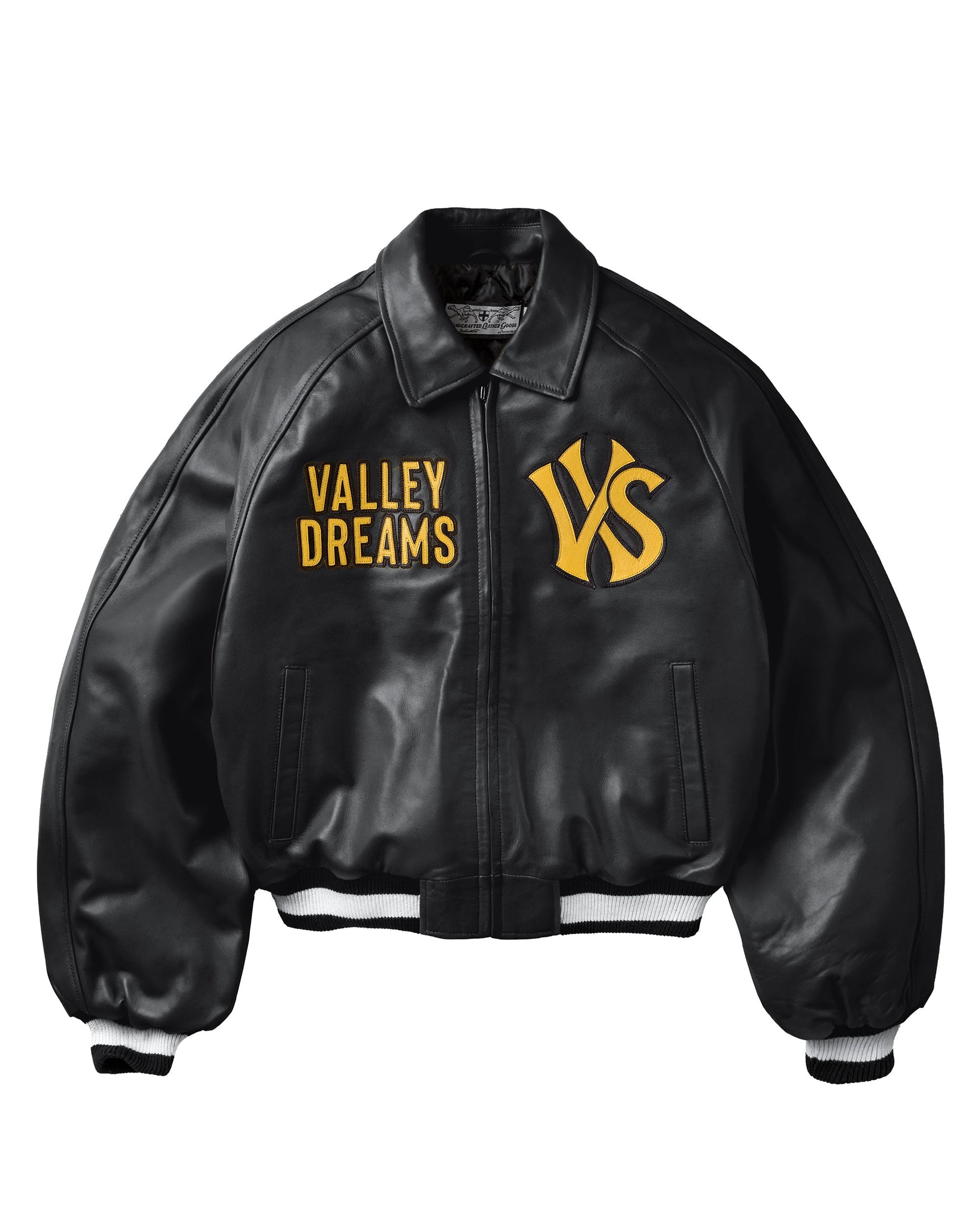 A black leather bomber jacket with yellow embroidered text reading 'VALLEY DREAMS' on the left chest and a large yellow 'VS' monogram on the right chest. The jacket features a front zipper, ribbed collar, cuffs, and hem with white stripes.